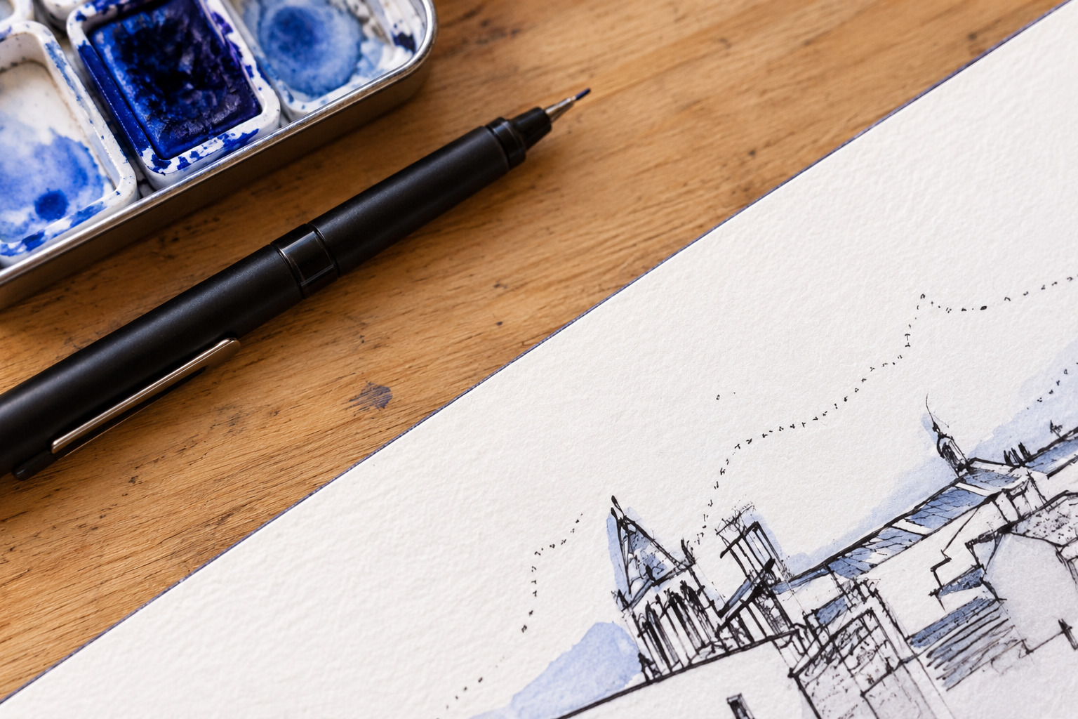 Detalle de un sketchbook abierto con dibujo a tinta negra, sombras de acuarela azul, un boli y una caja de acuarelas sobre una mesa de madera